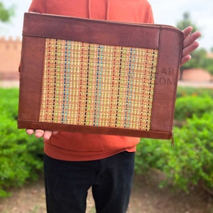 May include: A brown leather laptop case with a woven, multi-colored fabric insert. The case has a zipper closure.