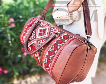 Puede incluir: Una bolsa de lona de cuero marrón con una sección central textil estampada. El textil presenta un diseño geométrico rojo, blanco y negro. La bolsa tiene una correa para el hombro y asas, y se lleva al hombro.