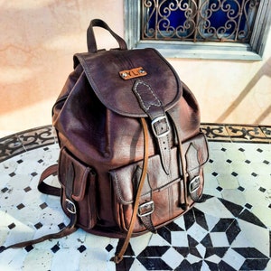 May include: A brown leather backpack with a flap closure and adjustable straps. The backpack has a small leather patch with the letters "YLI" on it.