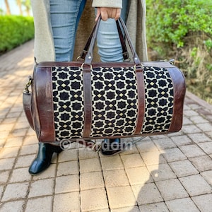 May include: A brown leather and patterned fabric duffel bag with a geometric design. The bag has a rectangular shape with a zipper closure, top handles, and a shoulder strap. The fabric is black with a white and gold geometric pattern.