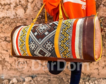 Puede incluir: Una bolsa de viaje de cuero marrón con ribetes amarillos y un patrón tejido colorido. La bolsa tiene un asa superior y una correa para el hombro.