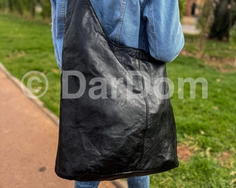 May include: A black leather shoulder bag with a long strap. The bag is worn over the shoulder, and the strap is visible. The bag is made of a textured leather material. The background shows a person wearing a denim jacket.