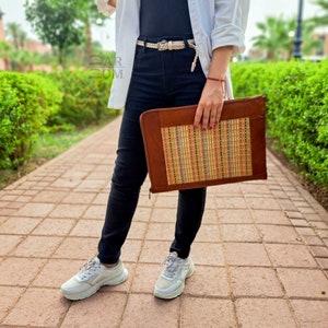 May include: A brown leather laptop sleeve with a woven pattern in shades of orange, yellow, and blue. The sleeve is being held by a person wearing black jeans and white sneakers.