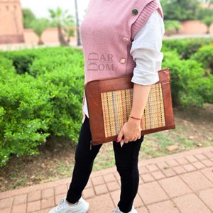 May include: A brown leather laptop sleeve with a woven colorful fabric panel. The sleeve has a zipper closure and is being held by a person.