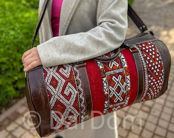 Puede incluir: Una bolsa de viaje de cuero marrón con un tejido estampado en rojo, blanco y naranja. La bolsa tiene una correa larga y un asa superior.