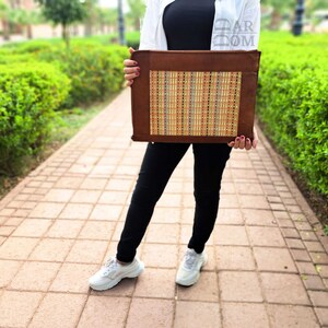 May include: A brown leather laptop case with a woven straw insert. The case is being held by a person wearing black pants and white sneakers.