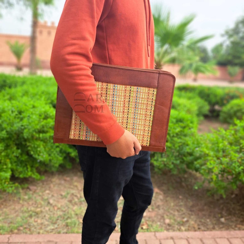 May include: A brown leather laptop sleeve with a woven straw front panel. The sleeve has a flap closure and is held by a person wearing a red sweater and black jeans.
