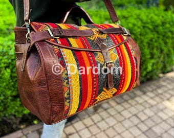 Op de afbeelding: Een bruine leren duffeltas met een kleurrijk gestreept stoffen paneel. De stof heeft verticale strepen in rood, oranje, geel en zwart. De tas heeft een schouderriem en gespsluitingen. De merknaam "Dardom" is zichtbaar.