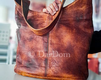 May include: A large, brown leather tote bag with a distressed finish. The bag has a wide, curved handle and a vertical seam down the center. The bag is held by a person's hand. The text "© DarDom" is visible.
