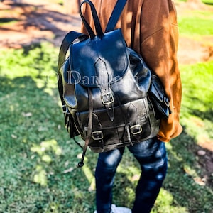 Pode incluir: Uma mochila de couro preto com vários bolsos e fivelas. A mochila tem um fecho de cordão e correias ajustáveis.