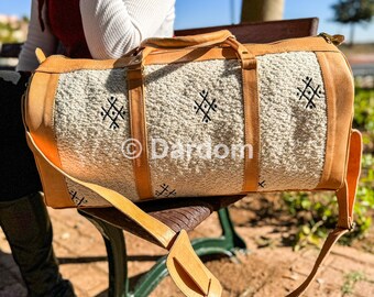 May include: A tan and cream-colored duffel bag with leather accents. The bag features a textured cream-colored center with black geometric designs. It has tan leather handles and a shoulder strap. The bag is sitting on a bench.