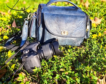 May include: A blue leather camera bag with a zipper and a black camera with a lens attached. The bag and camera are sitting on a bed of green grass with yellow flowers.