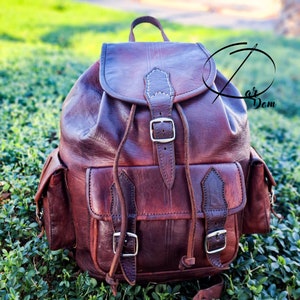 Puede incluir: Una mochila de cuero marrón con cierre de solapa, dos bolsillos delanteros y correas ajustables. La mochila tiene hebillas de color plateado y detalles de costura blancos. La bolsa está sobre un lecho de follaje verde.