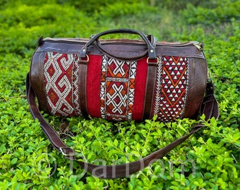 Puede incluir: Una bolsa de lona de cuero marrón con un panel central rojo con bordados geométricos en blanco, naranja y marrón. La bolsa tiene una correa para el hombro y asas superiores. La bolsa está colocada sobre un fondo de follaje verde.