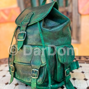 Może przedstawiać: Zielony skórzany plecak z wieloma kieszeniami i klamrami. Plecak ma zapięcie na klapę i regulowane paski.