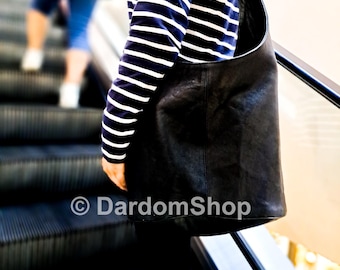 May include: A black leather shoulder bag with a curved top and a single strap. The bag is worn over the shoulder. The person in the image is wearing a blue and white striped long-sleeve shirt. The image is taken on an escalator.
