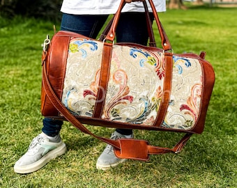 Puede incluir: Una bolsa de lona de cuero marrón con un panel central color crema con un diseño floral colorido. La bolsa tiene asas de cuero marrón y una correa para el hombro. La bolsa es sostenida por una persona de pie sobre hierba verde.