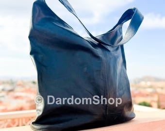 May include: A black leather shoulder bag with a single strap. The bag is displayed outdoors against a backdrop of a blue sky and a cityscape. The bag has a smooth texture and a simple design. The text "DardomShop" is visible on the bag.