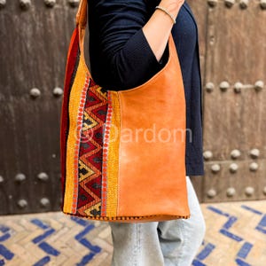 Könnte beinhalten: Eine hellbraune Leder-Umhängetasche mit einem farbenfrohen Webstoffpaneel. Die Tasche hat ein großes, offenes Design mit einem einzelnen Schulterriemen. Das gewebte Panel zeigt rote, gelbe und schwarze geometrische Muster. Das Wort "Dardom" ist auf der Tasche aufgedruckt.
