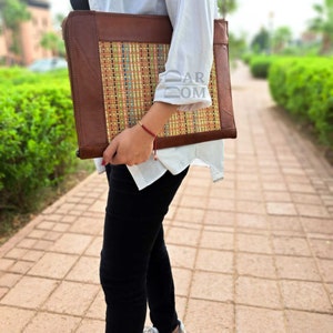 May include: A brown leather laptop sleeve with a woven colorful pattern. The sleeve is held by a person wearing a white shirt and black pants.