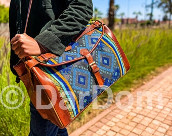 May include: A brown leather messenger bag with a blue, yellow, and gray patterned fabric flap. The bag has a long strap and a buckle closure.