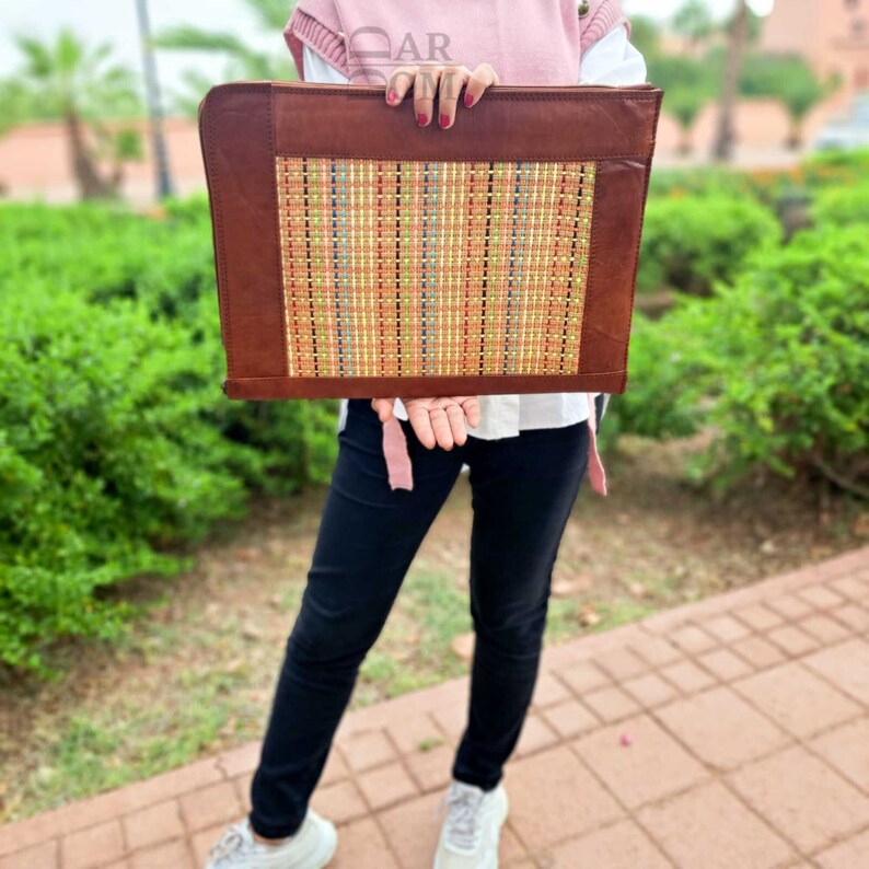 May include: A brown leather laptop sleeve with a woven pattern in shades of orange, yellow, and blue. The sleeve is held in front of a person wearing black pants and a pink shirt.
