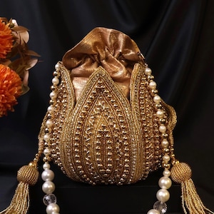 Puede incluir: Un bolso dorado con cordón y cuentas con un diseño floral. El bolso está adornado con perlas y borlas doradas. El fondo es negro, con flores naranjas en la esquina superior izquierda.