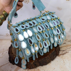 May include: A light blue handbag with a suede-like texture, adorned with teardrop-shaped embellishments in various colors. The bag features a matching handle and gold-colored accents. The design is intricate and eye-catching, suitable for special occasions.