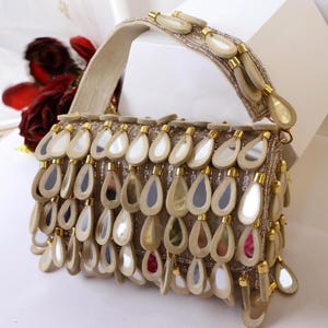 Könnte beinhalten: Eine beige Handtasche mit einem strukturierten Griff und einem Körper, der mit zahlreichen hängenden, tropfenförmigen Spiegelakzenten verziert ist. Die Oberfläche ist mit Reihen dieser reflektierenden, metallisch eingefassten Elemente bedeckt.