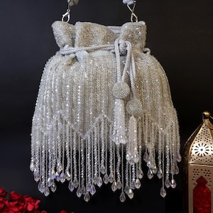 Könnte beinhalten: Eine silberne, mit Perlen besetzte Handtasche mit Kordelzug und Schulterriemen. Die Tasche ist mit hängenden Kristallperlen und silbernen Quasten verziert. Ein rotes Blumenarrangement und eine goldene Laterne befinden sich im Hintergrund.
