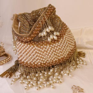 Puede incluir: Un bolso con cordón de cuentas doradas y blancas. El bolso está cubierto de pequeñas cuentas redondas, con una franja de perlas y borlas doradas. La parte superior del bolso tiene un ribete de cuentas doradas y un cierre de cordón. Se ve una pulsera dorada a juego.