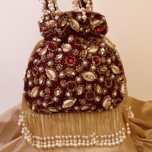 May include: A burgundy beaded handbag with gold and pearl accents. The bag features a rounded shape, embellished with red gems, gold sequins, and white shell-shaped details. Pearl strands form the handle and a decorative fringe.