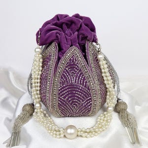 Puede incluir: Un bolso morado de terciopelo con cordón, intrincado trabajo de cuentas plateadas y una correa de perlas y cuentas. El bolso presenta un diseño decorativo y está adornado con borlas. El bolso está sobre un fondo blanco.