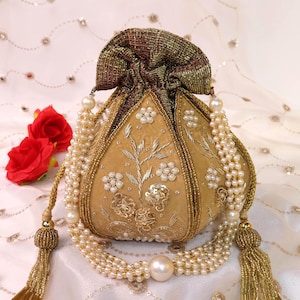 Puede incluir: Un bolso tipo bolsa dorado y con cuentas con cierre de cordón. El bolso presenta bordados florales y está adornado con hilos de perlas y borlas. El fondo incluye rosas rojas y una tela blanca estampada.