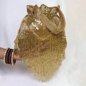 May include: A gold beaded drawstring bag with a matching strap and tassel. The bag is embellished with hanging beads and a decorative flap. A person's arm is visible, adorned with maroon and gold bangles.