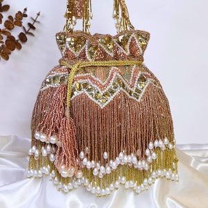 Puede incluir: Un bolso de mano con cuentas en tonos oro rosa, dorado y blanco. El bolso presenta un cierre de cordón, una borla y una franja de perlas. El asa también está adornada con cuentas. Ideal para ocasiones especiales.