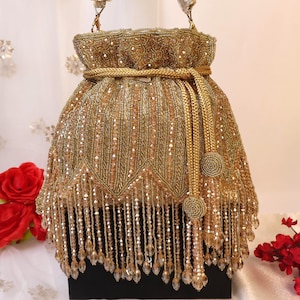 Könnte beinhalten: Eine goldfarbene, mit Perlen besetzte Handtasche mit Kordelzugverschluss und langem Schulterriemen. Die Tasche weist aufwendige Perlenarbeiten und eine Fransenbordüre aus hängenden Perlen am unteren Rand auf. Die Tasche steht auf einer schwarzen Oberfläche.