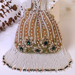 May include: An ornate, beaded handbag with a gold-colored interior. The bag is covered in white, peach, and clear beads, with a fringe of white beads at the bottom. Green and gold embellishments with emerald-colored stones adorn the front.