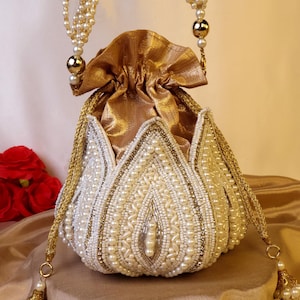 Puede incluir: Un bolso de mano blanco con cuentas, con cierre de cordón dorado y asa de cuentas doradas. El bolso está adornado con perlas y detalles dorados, y tiene una borla de perlas. El bolso está sobre una superficie de satén dorado.