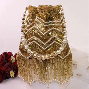May include: A gold beaded drawstring bag with a chevron pattern. The bag features white pearl accents and a beaded fringe. The handle is made of pearls and gold beads. The bag is on a white surface.