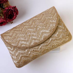 Op de afbeelding: Een goudkleurige clutch met een klepsluiting. De clutch is bedekt met kleine, glinsterende kralen in een zigzagpatroon. De randen zijn ook afgezet met kralen. Een paar rode rozen op de achtergrond. Een stijlvol accessoire.