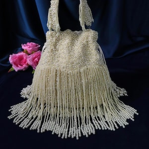 May include: Cream-coloured beaded handbag with a rounded shape and a long, beaded fringe. The bag has a curved handle and is adorned with numerous small beads. Pink roses are in the background.