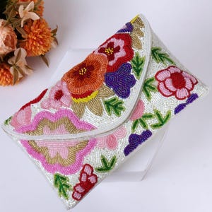 以下が含まれることがあります： カラフルな花の模様が施された、白いビーズのクラッチバッグ。花は赤、オレンジ、ピンク、紫、金色の色合いで、緑の葉が付いています。クラッチバッグはフラップ開閉式で、特別な機会に適しています。