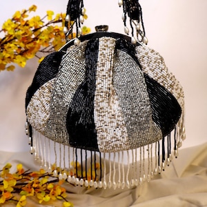 Peut inclure: Un sac à main rond en perles noires, blanches et argentées avec une longue bandoulière en perles. Le sac a une frange de perles blanches autour du bas.