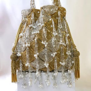 May include: A gold and silver beaded handbag with a structured shape. The bag features a scalloped design, pearl accents, and dangling beaded strands. Gold tassels hang from the sides. The handles are gold.