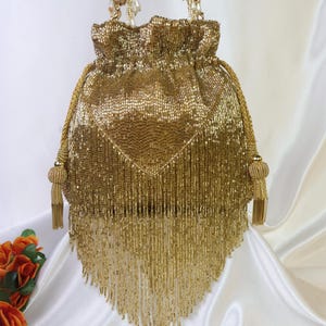 May include: A gold beaded drawstring bag with a tassel fringe. The bag has a gathered top and beaded handles. The bag is a shimmering gold color and is likely a formal accessory.