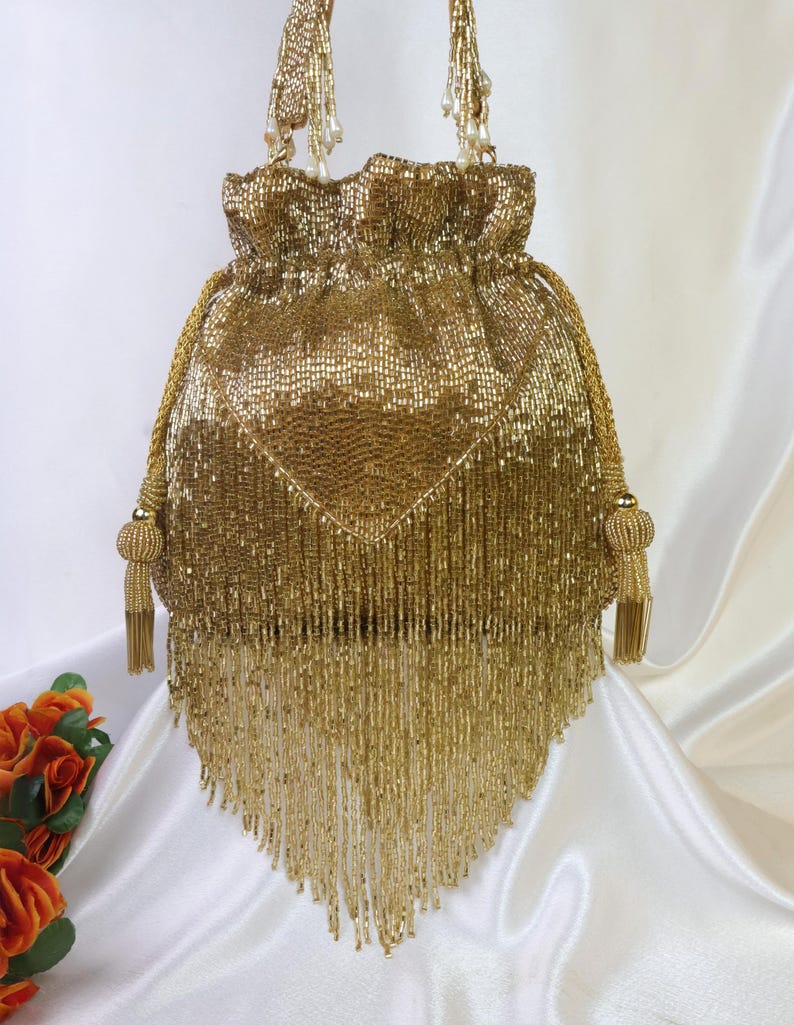 May include: A gold beaded drawstring bag with a tassel fringe. The bag has a gathered top and beaded handles. The bag is a shimmering gold color and is likely a formal accessory.