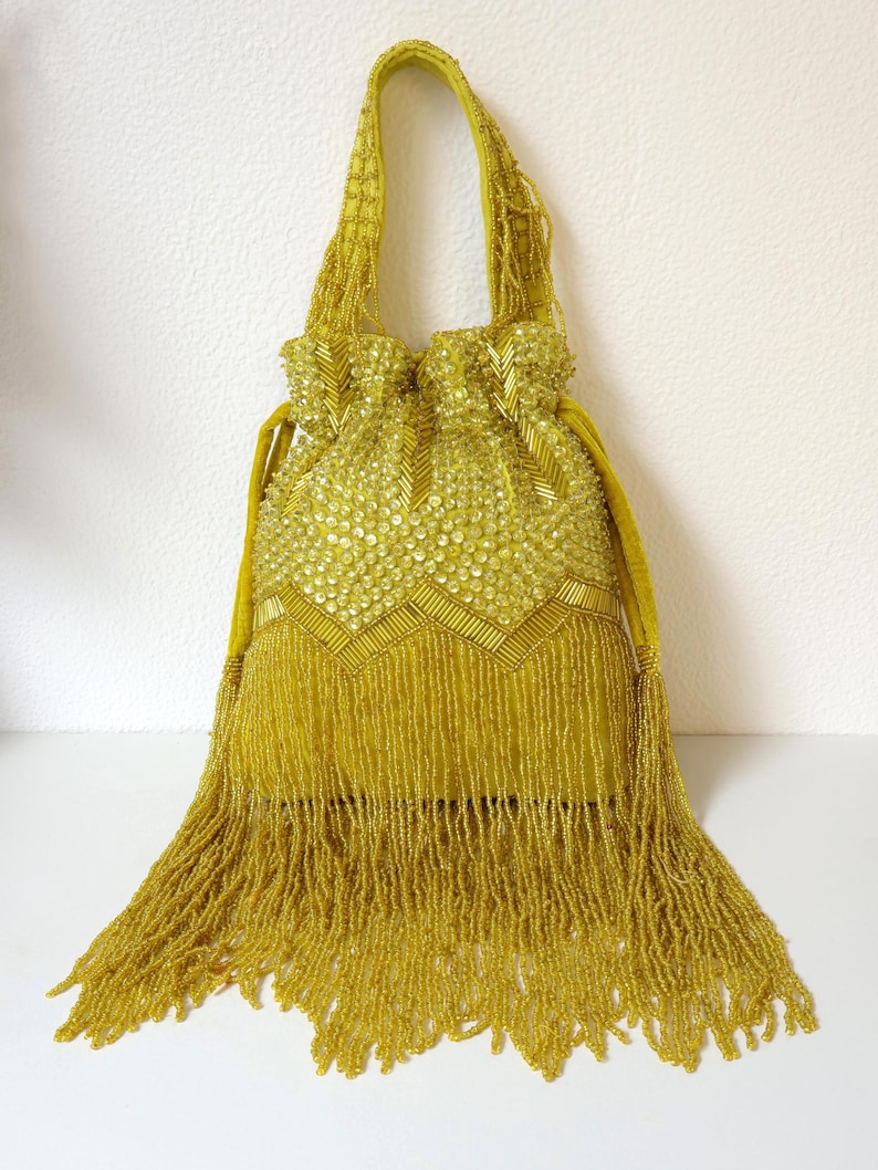May include: A chartreuse drawstring handbag featuring an intricate exterior covered in shimmering sequins and glass beads. The bag includes a matching beaded handle, a chevron-patterned upper section, and a long, dense fringe of beads at the base.