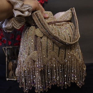 May include: A gold beaded handbag with a drawstring closure and a handle. The bag is embellished with rows of beads and dangling crystal-like accents. The bag is held by a person wearing a beige scrunchie.