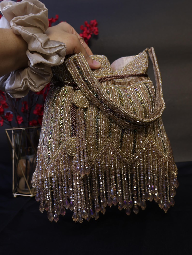 May include: A gold beaded handbag with a drawstring closure and a handle. The bag is embellished with rows of beads and dangling crystal-like accents. The bag is held by a person wearing a beige scrunchie.